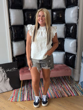 Load image into Gallery viewer, A woman standing in front of a black and white decorative wall, wearing a white eyelet top with ruffle sleeves and denim shorts, with sneakers on her feet.

